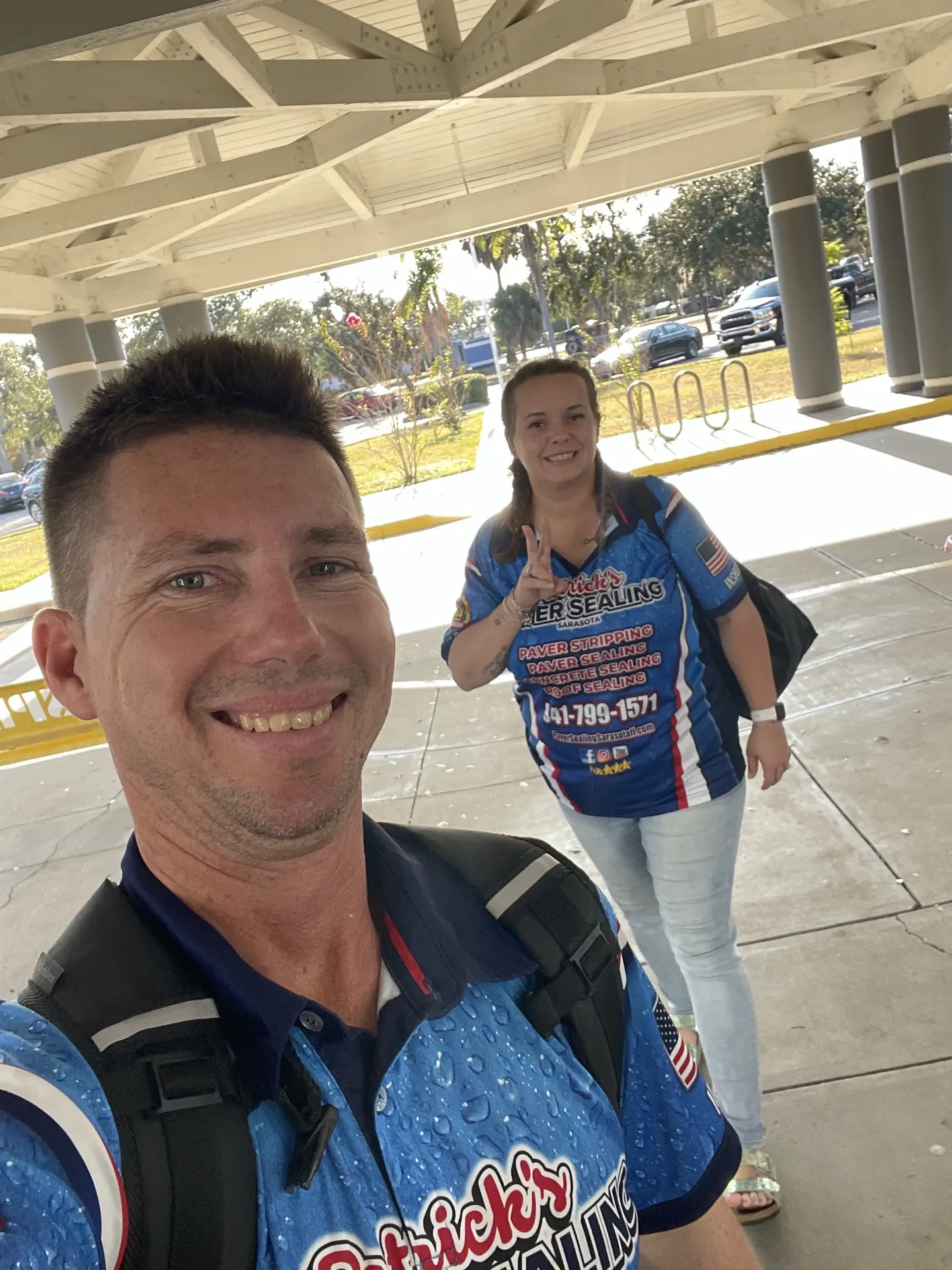 A smiling man taking a selfie under a covered entry, with a woman in a matching blue jersey waving behind him