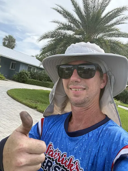 Smiling team member in a brim hat and sunglasses giving a thumbs up in front of a paver driveway, in a blue branded shirt