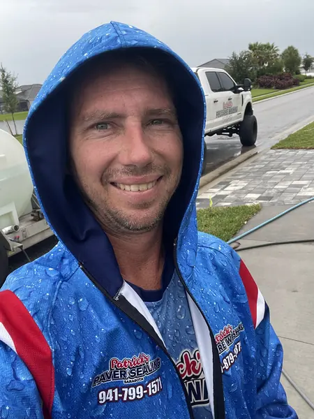 Smiling team member in a blue hooded branded jacket in light rain, a white pickup, water tank and paver driveway behind