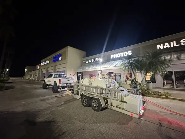 Night view of a shopping plaza with a pressure washing truck and trailer parked in the lot and an orange hose running out