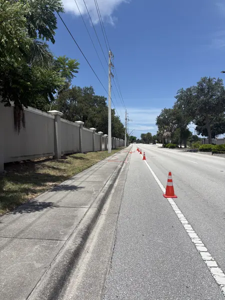 Sidewalk and road beside a long tall perimeter wall, with traffic cones and an orange hose laid along the curb