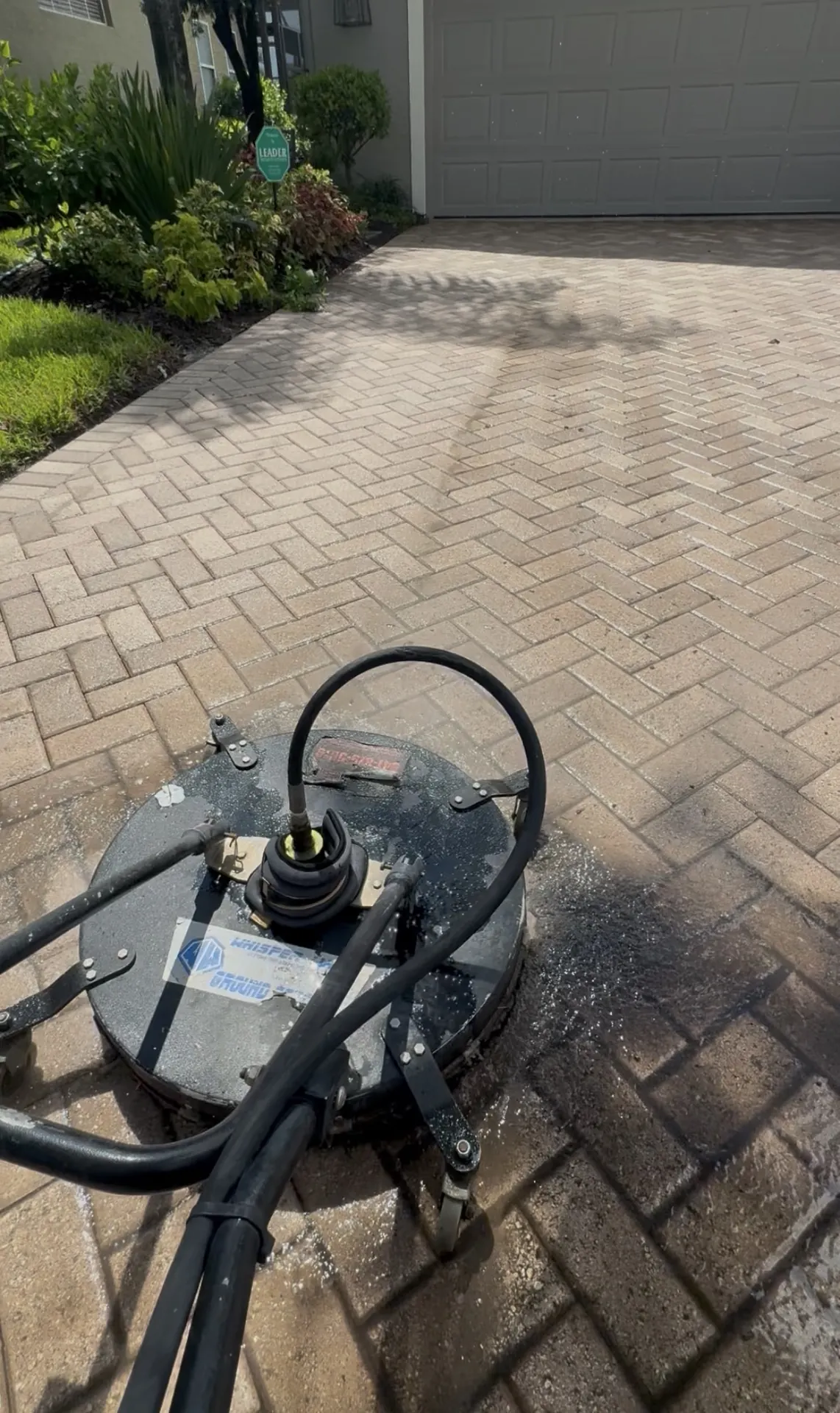 Gray surface cleaner disc on a paver walkway beside landscaping, with a lighter cleaned strip behind it