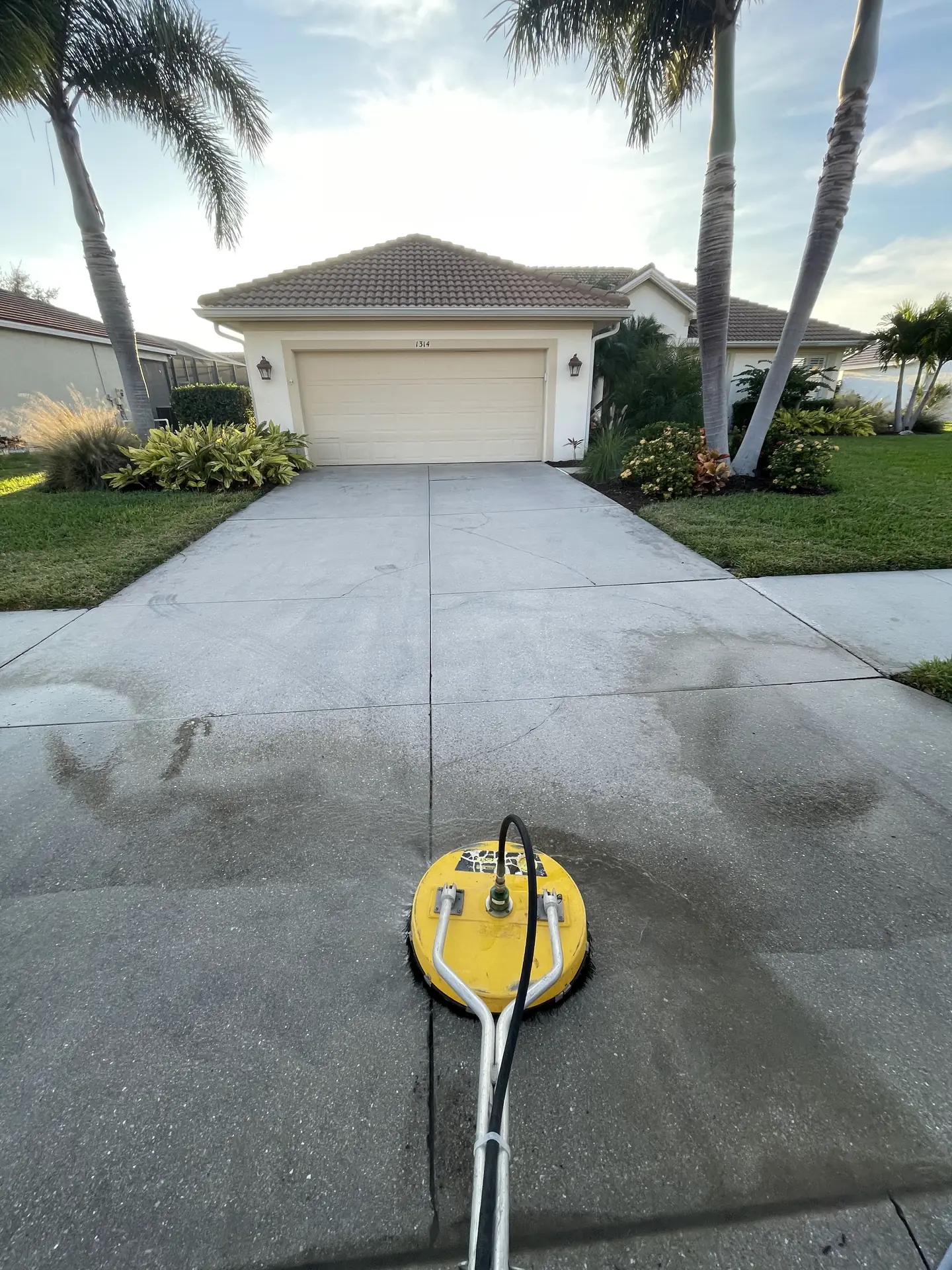 Yellow surface cleaner on a concrete driveway in front of a tan garage, half the driveway already washed