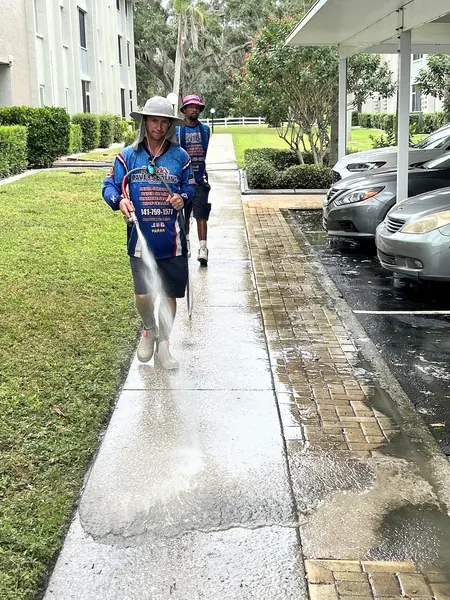 Worker in a wide brim hat spraying a wet concrete sidewalk beside brick pavers while a second worker follows