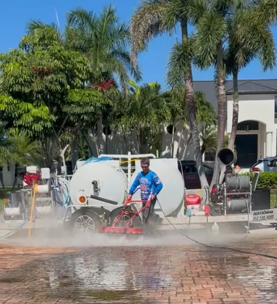 Worker operating a red surface cleaner on a paver drive with mist rising and the trailer rig behind him