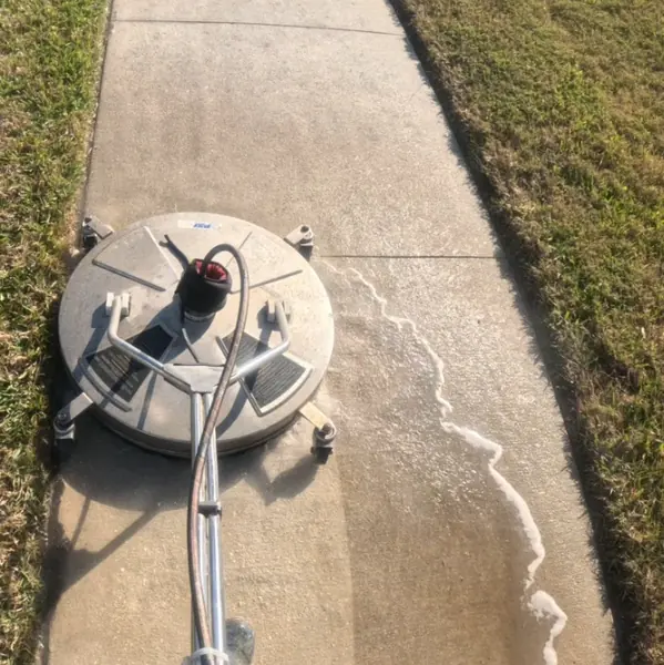 Gray rotary surface cleaner on a concrete sidewalk with a foam edge and a lighter cleaned strip behind it
