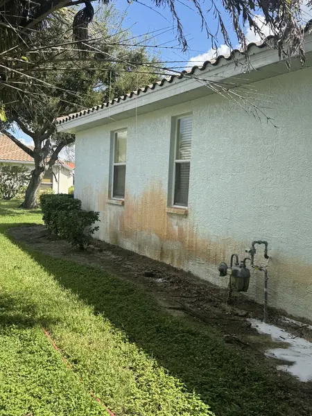 Stucco house side wall with orange rust stains below two windows, with a gas meter and mulch bed beside it