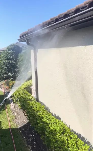 Water spraying up a cream stucco wall to the roof edge, where a dark gutter runs along the eave