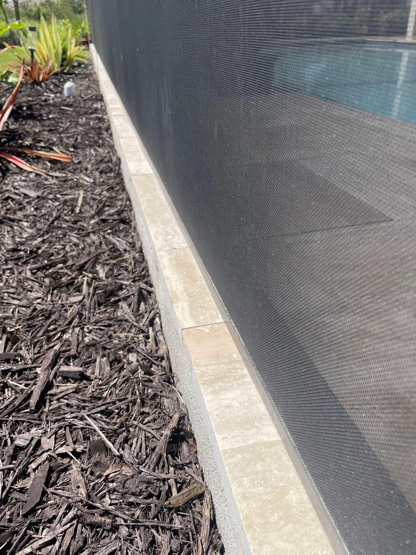 Pale travertine pool deck tiles beside mulch and a screen, with a dull dusty film