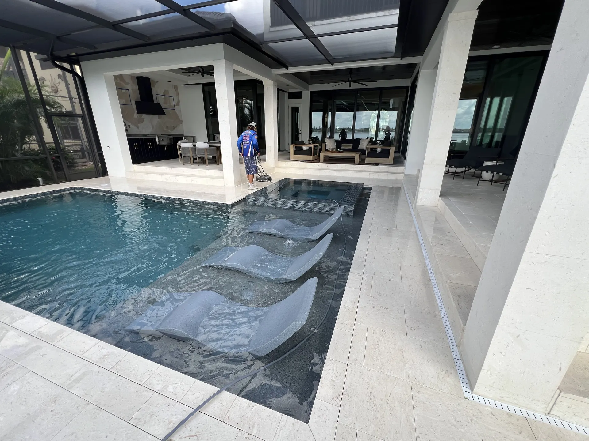 Worker guiding a surface cleaner beside a spa on a pale stone pool deck, covered lanai seating behind