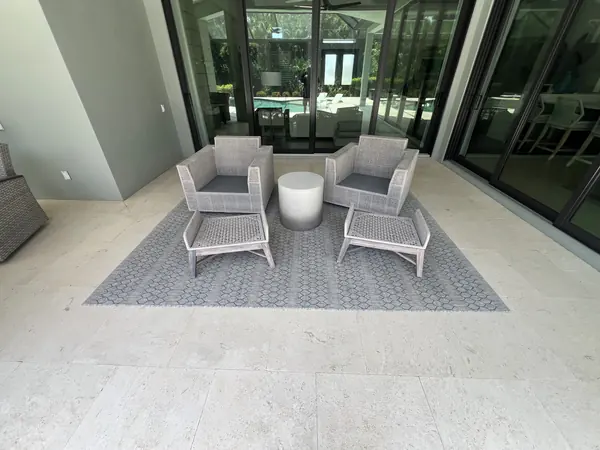 Pale stone floor of an outdoor living area with grey wicker chairs on a rug, glass doors and pool in the background