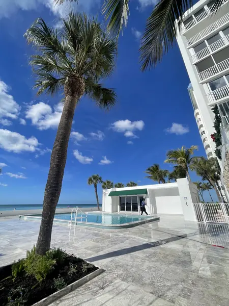 Beachfront condo pool deck with grey stone paving, a tall palm and a worker in the distance pulling a hose