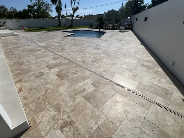Large travertine pool deck with a long drain channel, white boundary wall and a pool and yard beyond