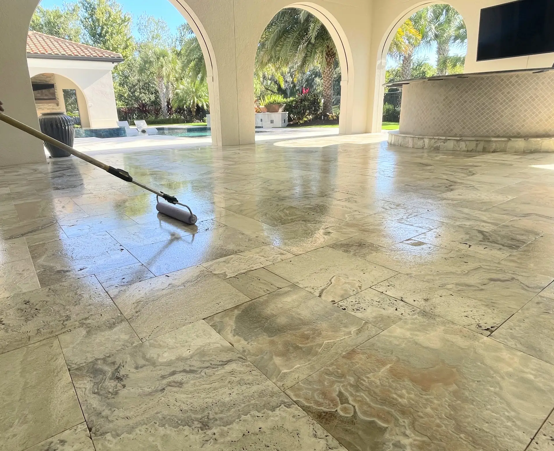 Pole mounted roller on a wet looking travertine floor in an arched covered patio with a pool in the background
