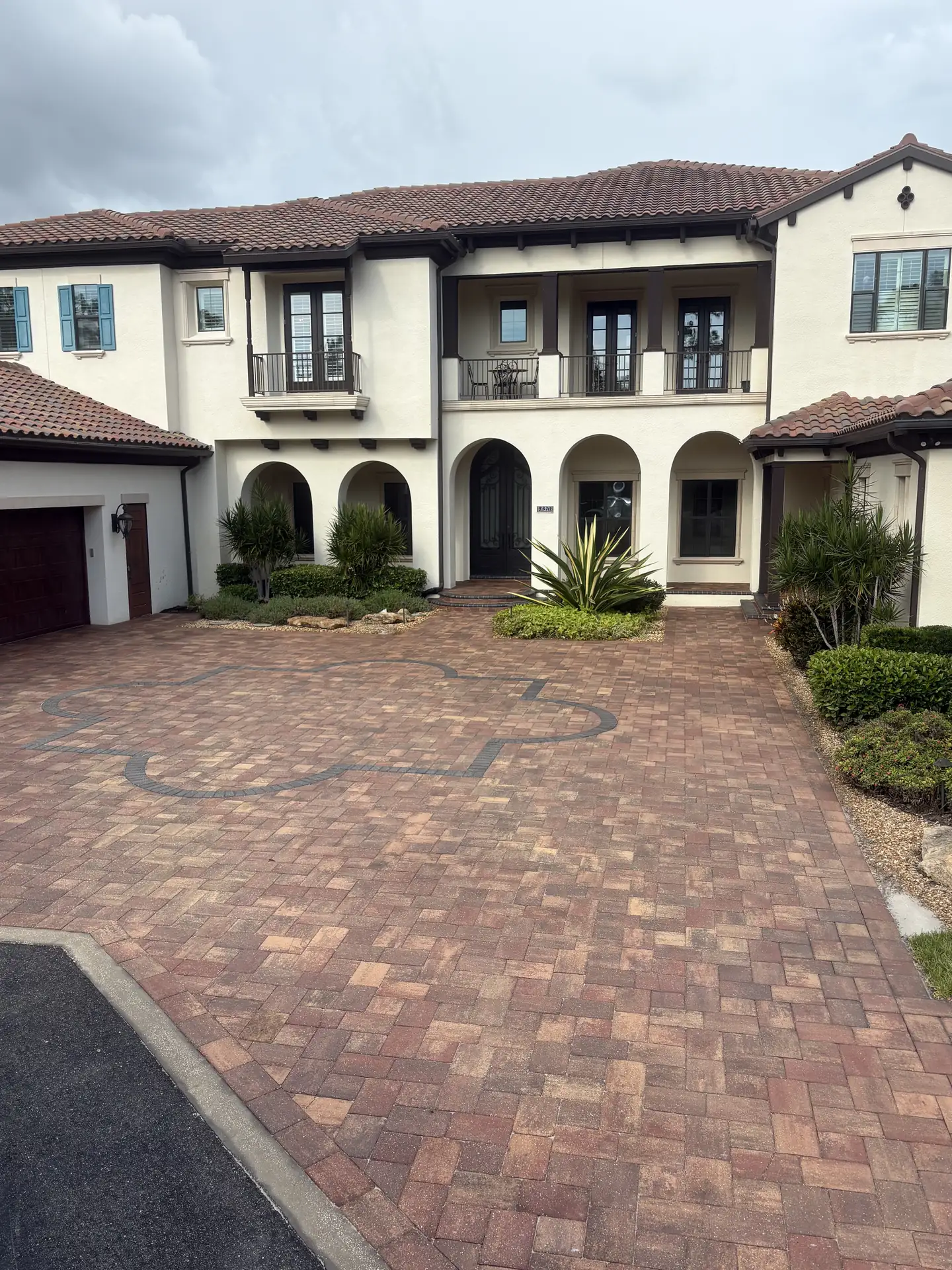 Brick paver driveway with a dark decorative inlay pattern in front of a two-story stucco home with a tile roof
