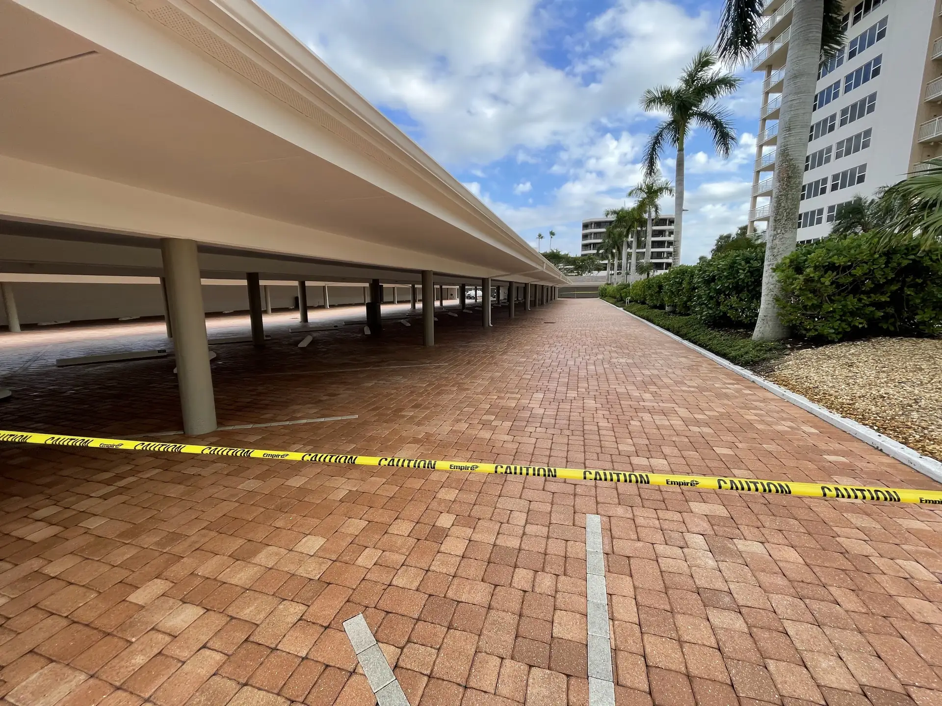 Long red brick paver drive beneath a multi-level covered parking structure beside a high-rise, with caution tape across it
