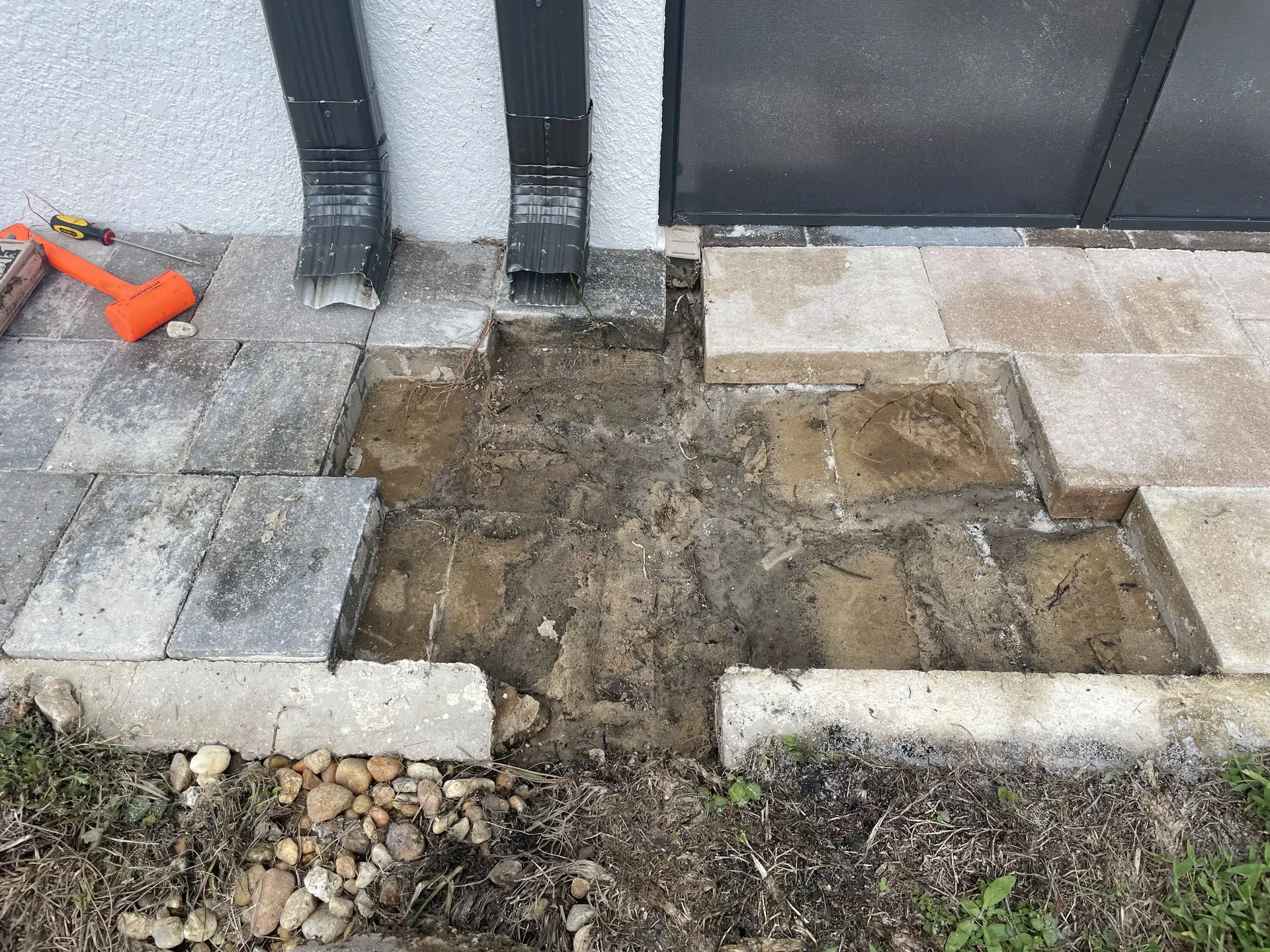 Section of pavers removed beside two downspouts, with bare earth base showing and an orange mallet on the surface