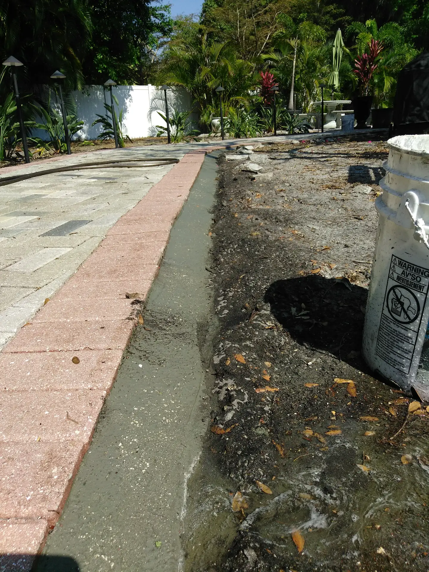 Brick paver border beside a fresh gray concrete edge strip, with a bucket and a hose, and garden lights in the background