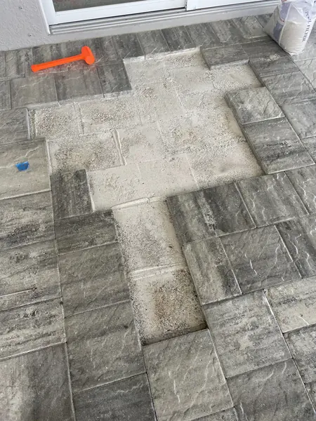 Gray pavers lifted beside a sliding door, bare bedding sand showing, with an orange mallet and a sand bag nearby