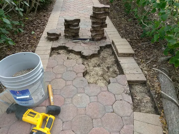Paver walkway section lifted and stacked, bare sand bed exposed, with a mallet, bucket, trowel and saw on the ground