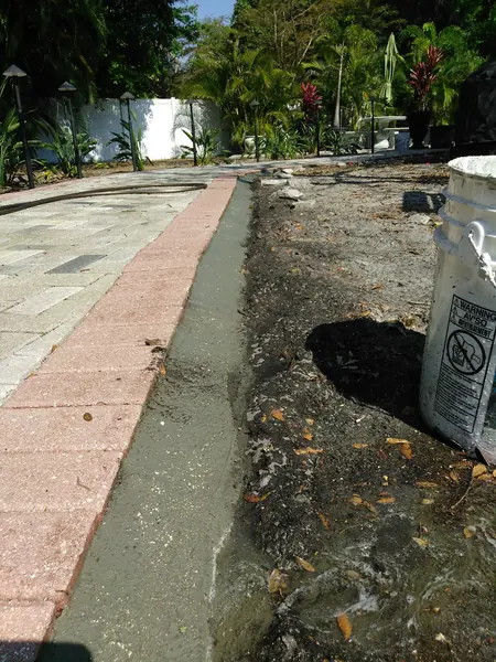Brick paver border beside a fresh gray concrete edge strip, with a bucket and a hose, and garden lights in the background