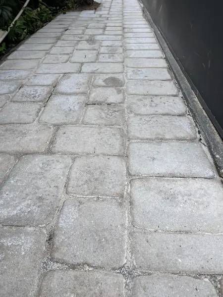 Weathered gray interlocking pavers along a walkway beside a dark wall, with a dull surface and chalky joints