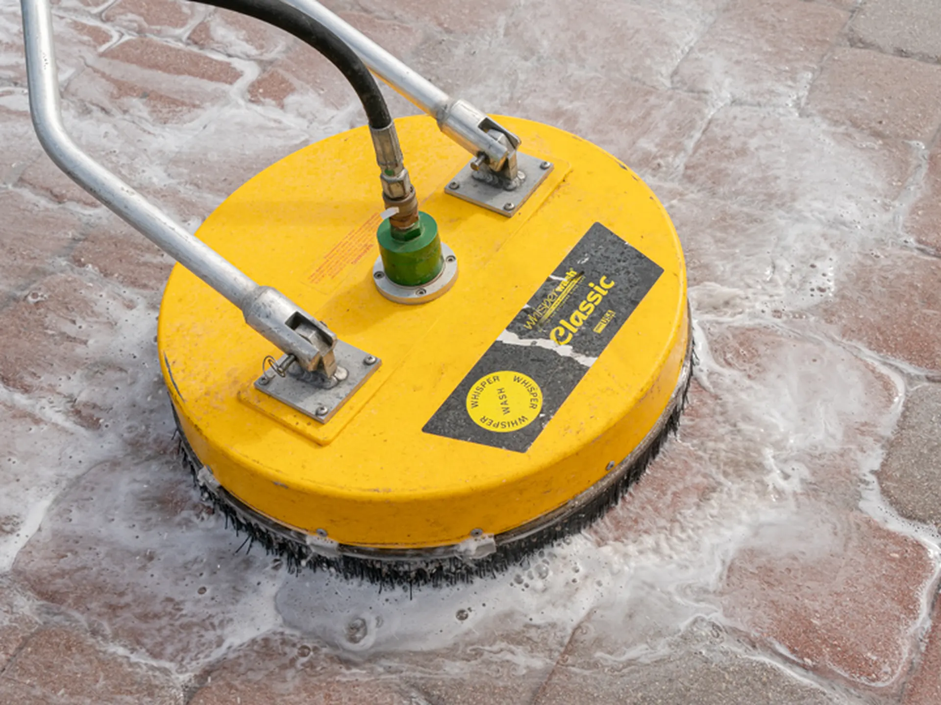 Yellow rotary surface cleaner scrubbing red brick pavers covered in white foam, close view