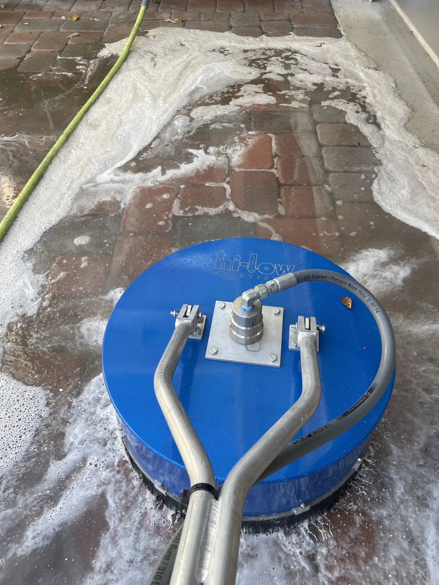 Blue rotary surface cleaner scrubbing brick pavers with thick white foam and a green hose in the frame