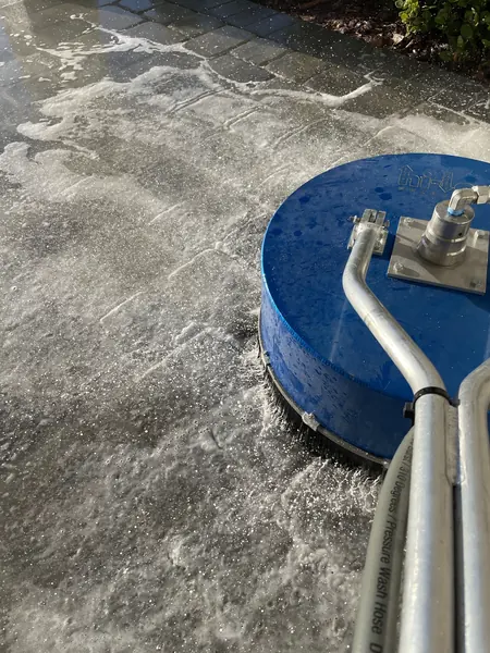 Blue rotary surface cleaner scrubbing a wet gray paver surface with foam gathering near a planting bed