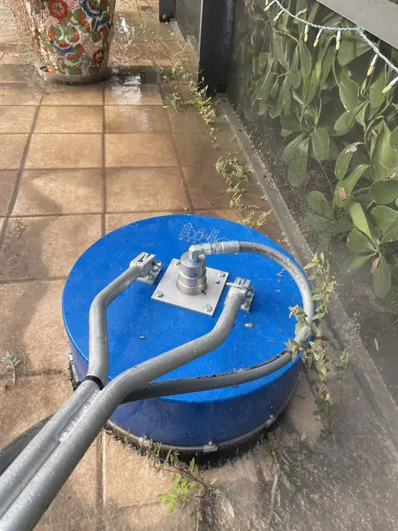 Blue rotary surface cleaner on square terracotta patio tiles beside a flower pot, a metal rail and a hedge