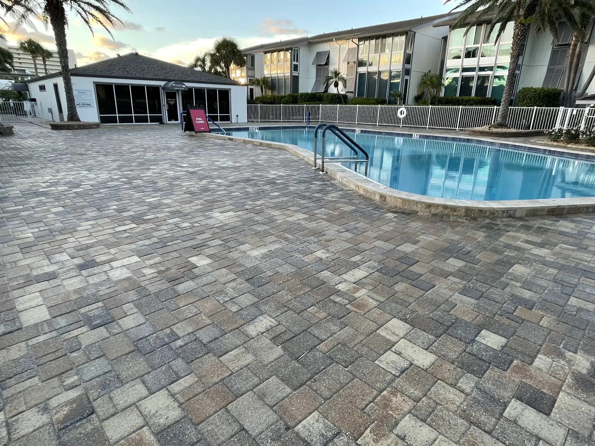 Grey paver pool deck with curved coping and a pool beside a multi unit building with a fence