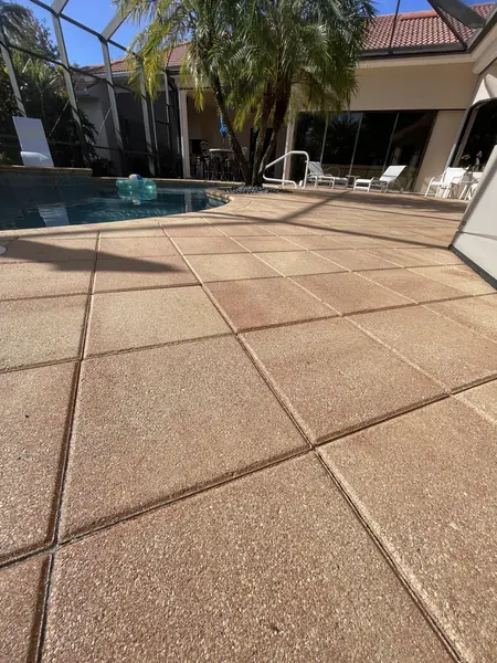 Tan textured concrete pool deck slabs with joints, pool in the background and a house and chairs behind