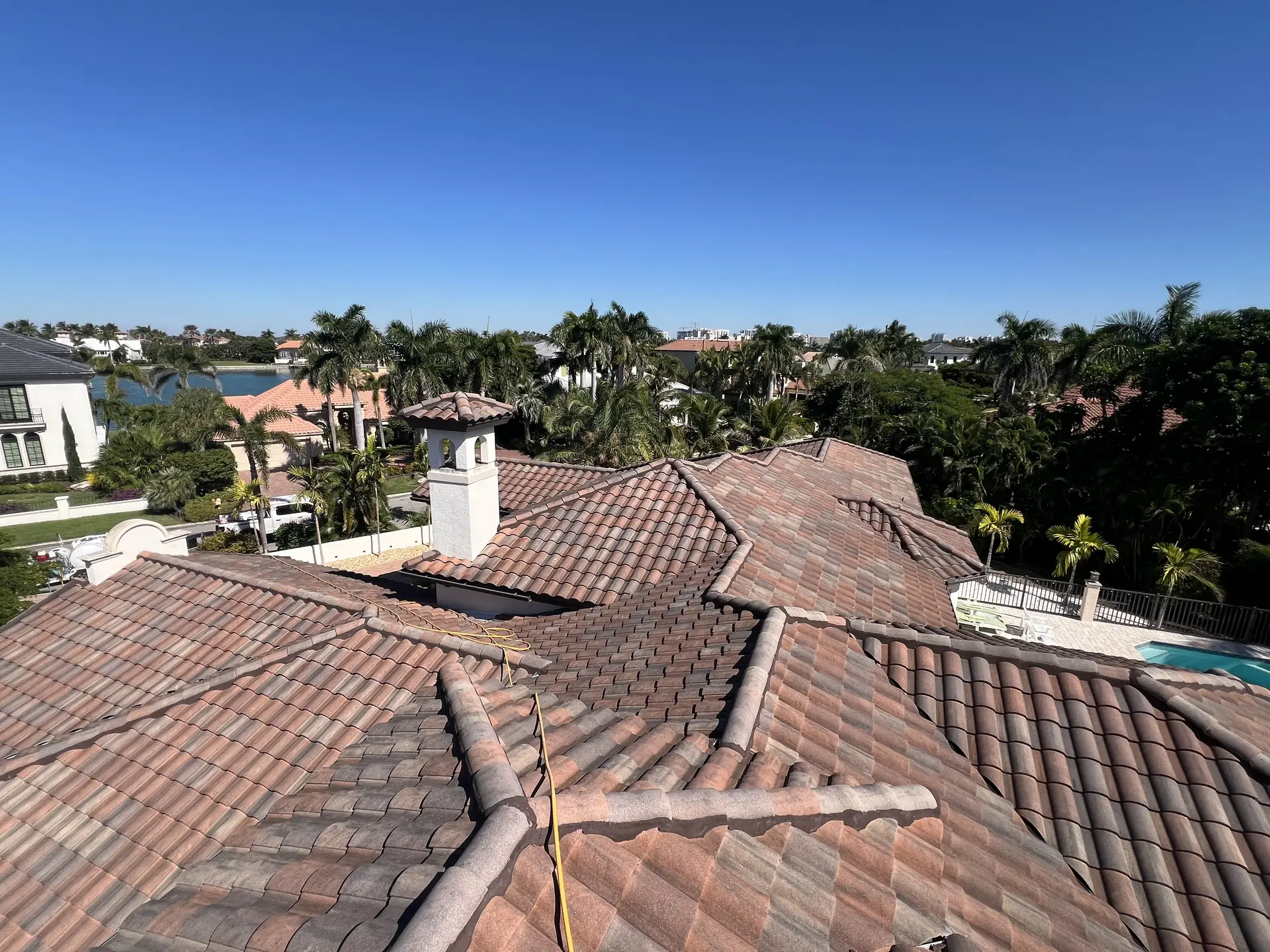 Aerial view over a clay tile roof with several ridges and a chimney, waterfront homes and blue sky behind
